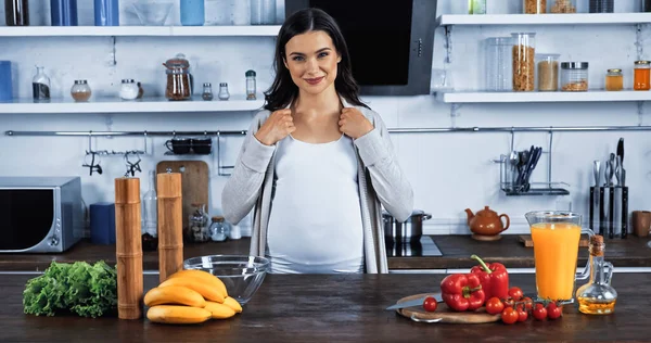 Улыбающаяся беременная женщина стоит рядом со свежими овощами, бананами и апельсиновым соком на кухне — стоковое фото