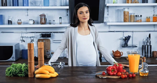Беременная женщина смотрит в камеру рядом со свежими овощами и апельсиновым соком — стоковое фото