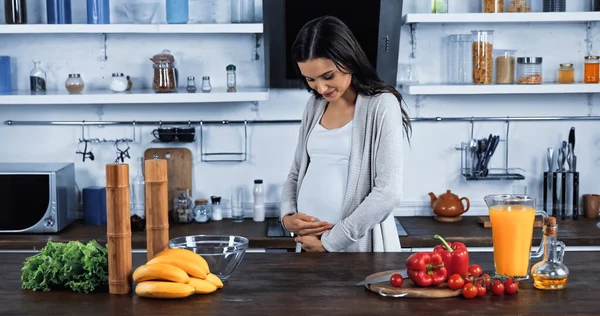 Junge Schwangere blickt auf Bauch neben frischem Essen auf Küchentisch — Stockfoto