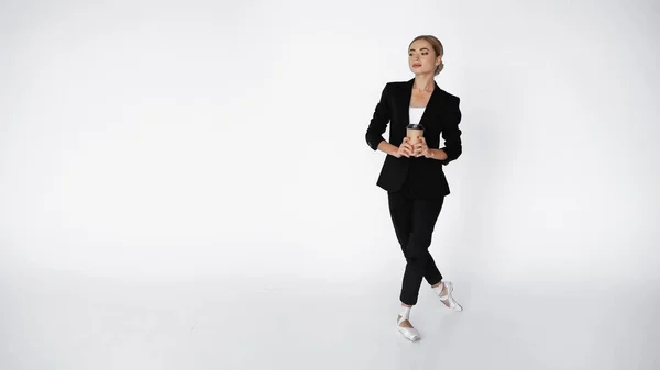 Elegante bailarina en traje y zapatos puntiagudos sosteniendo café para llevar mientras camina sobre blanco - foto de stock