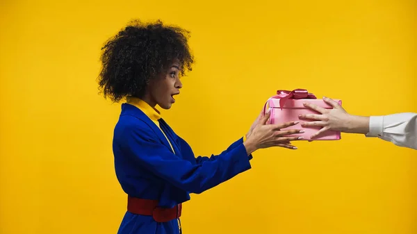 Vista lateral de la mujer afroamericana asombrada tomando caja de regalo aislada en amarillo - foto de stock
