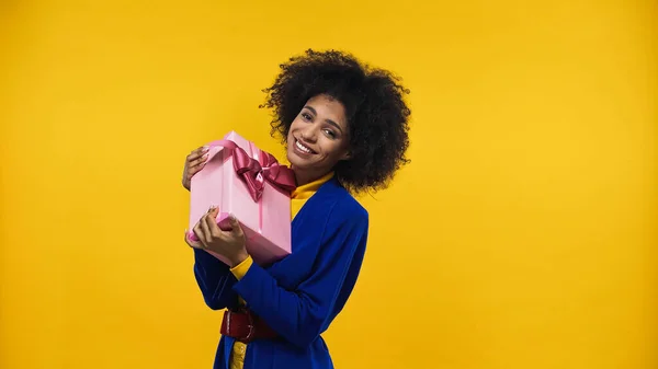 Happy african american woman holding pink present isolated on yellow — Stock Photo