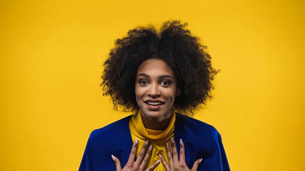 Lächelnde afrikanisch-amerikanische Frau mit Händen in Brustnähe, die auf gelb isoliert in die Kamera blickt — Stockfoto