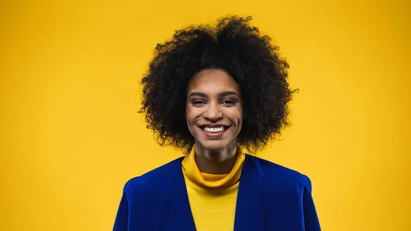 Mujer afroamericana positiva en blazer azul mirando la cámara aislada en amarillo - foto de stock