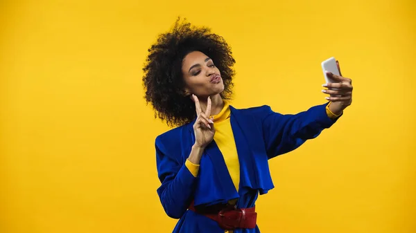 Femme afro-américaine montrant signe de paix pendant selfie isolé sur jaune — Photo de stock