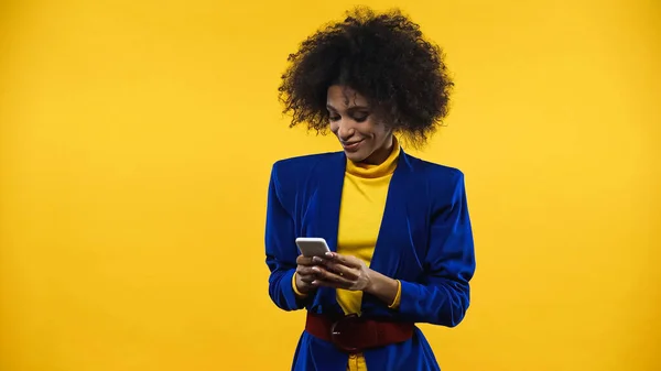 Femme afro-américaine souriante en veste bleue en utilisant un smartphone isolé sur jaune — Photo de stock