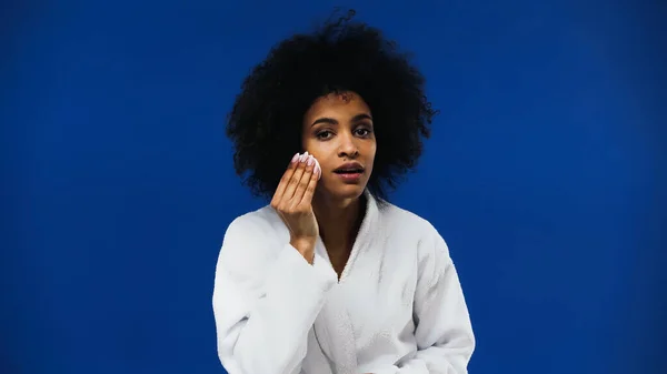 African american woman in white bathrobe using cotton pad on face isolated on blue — Stock Photo