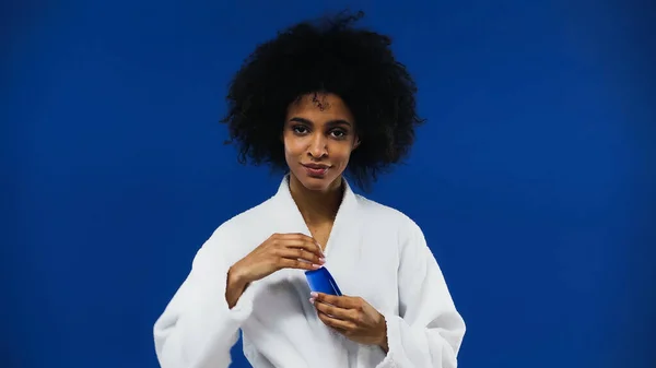 Femme afro-américaine en peignoir tenant lotion isolée sur bleu — Photo de stock