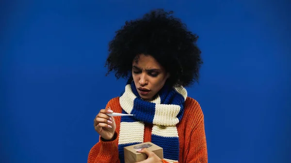 Ill femme afro-américaine tenant thermomètre et boîte isolée sur bleu — Photo de stock