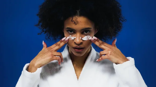 Mujer afroamericana en albornoz aplicando crema facial aislada en azul - foto de stock