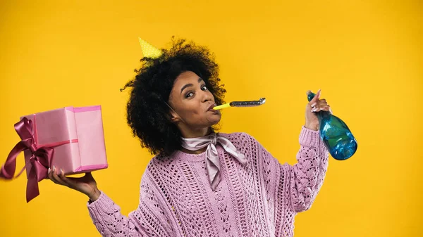 Femme afro-américaine en bonnet de fête tenant présent et bouteille de champagne isolé sur jaune — Photo de stock