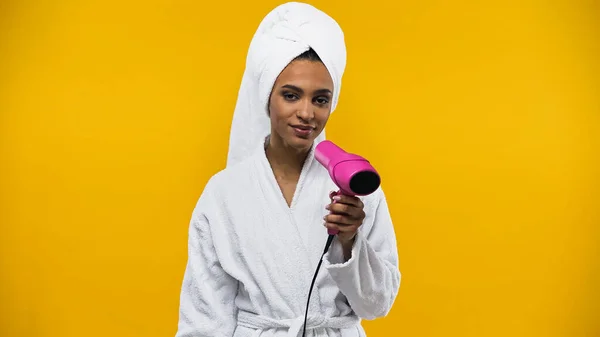 African american woman in bathrobe holding hairdryer isolated on yellow — Stock Photo