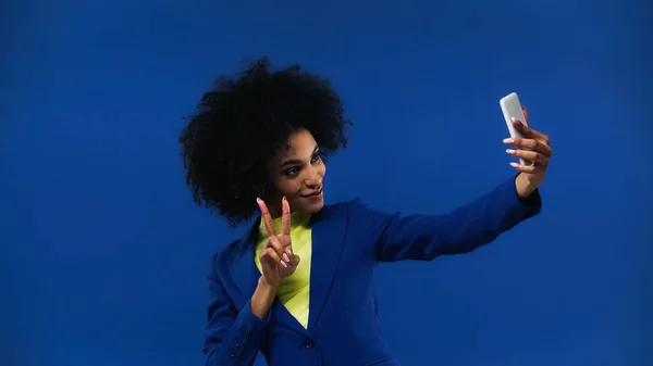 Femme afro-américaine souriante montrant un signe de paix et prenant selfie isolé sur bleu — Photo de stock