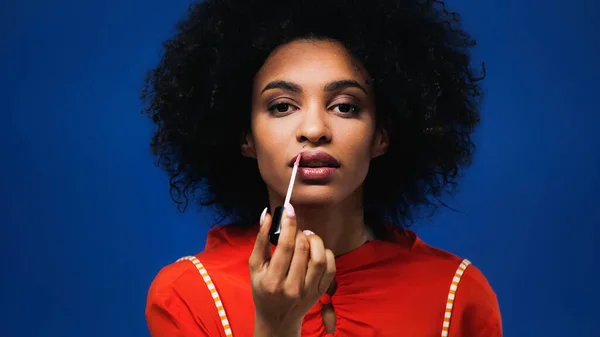 Jeune femme afro-américaine appliquant gloss lèvres isolées sur bleu — Photo de stock