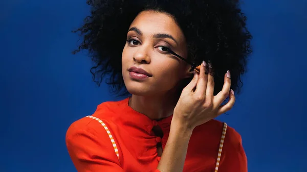 Mujer afroamericana aplicando rímel aislado sobre azul - foto de stock