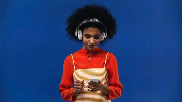 Donna afroamericana utilizzando cuffie e smartphone isolati su blu — Foto stock