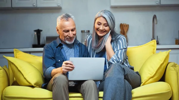 Веселая межрасовая пара сидит дома на диване с ноутбуком — стоковое фото