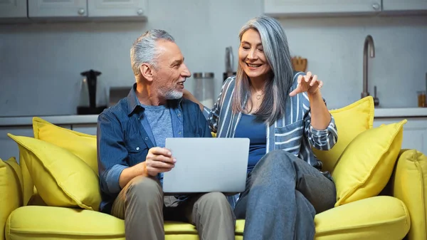 Cheerful asian woman gesturing while talking to husband near laptop at home — Stock Photo