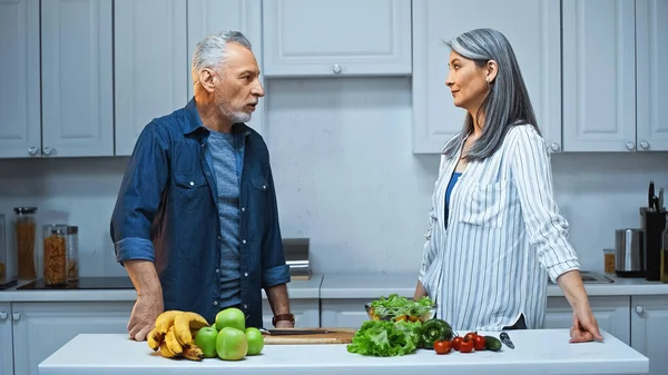 Casal multicultural cabelos grisalhos olhando uns para os outros, enquanto de pé perto de frutas e legumes na cozinha — Fotografia de Stock