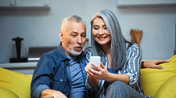 Веселая азиатская женщина, использующая мобильный телефон рядом с изумленным пожилым мужем — стоковое фото