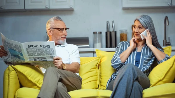Älterer Mann mit Zeitung schaut asiatische Frau an, die auf Smartphone spricht — Stockfoto