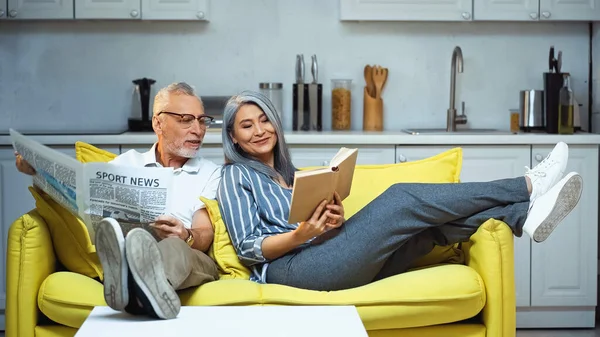 Sonriente asiático mujer leyendo libro cerca senior marido con periódico - foto de stock