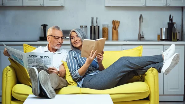 Cheerful asian woman showing book to elderly husband holding newspaper — Stock Photo