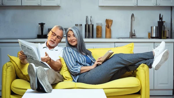 Senior man pointing at newspaper near asian wife sitting on sofa with book — Stock Photo