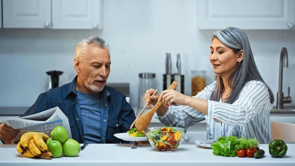 Senior asian woman mixing salad near husband with newspaper in kitchen — Stock Photo