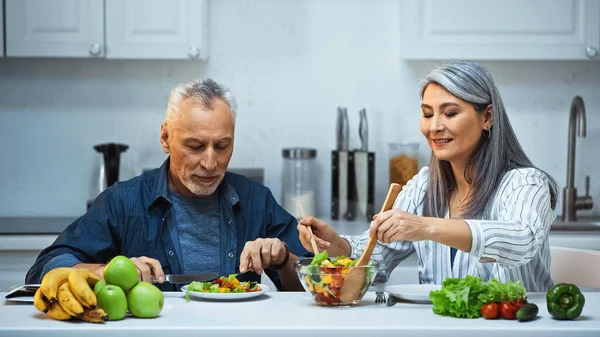 Smiling asian woman mixing salad near elderly husband having breakfast in kitchen — Stock Photo