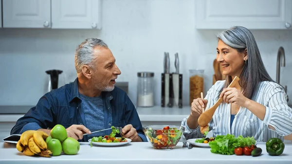 Happy senior interracial couple talking during breakfast in kitchen — Stock Photo