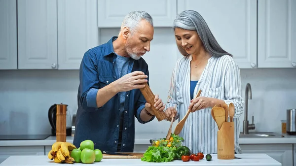 Homem sênior segurando moinho de especiarias perto feliz asiático esposa preparando salada para pequeno-almoço — Fotografia de Stock