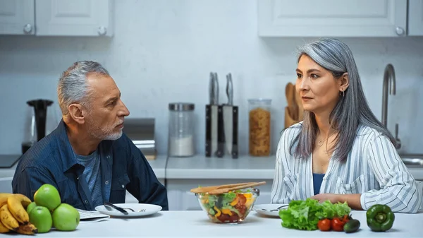 Pareja interracial de ancianos mirándose durante el desayuno en la cocina - foto de stock