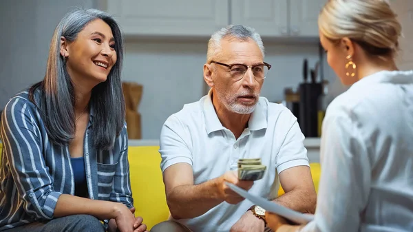 Asian woman smiling near elderly husband giving money to insurance agent on blurred foreground — Stock Photo