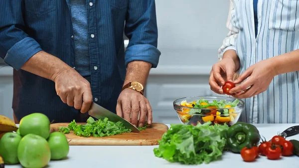 Vista recortada de pareja de ancianos preparando ensalada fresca en la cocina - foto de stock
