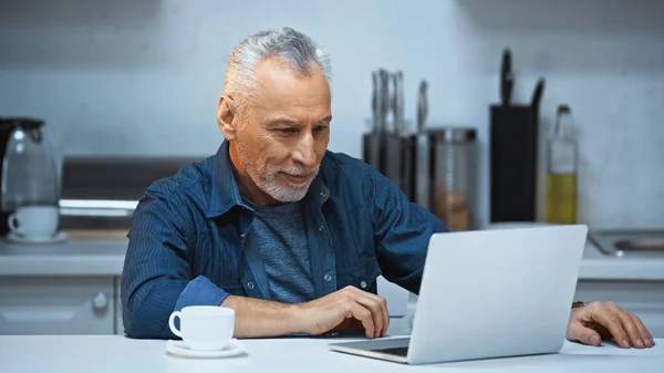 Freelancer de pelo gris que trabaja cerca del ordenador portátil y la taza de café en la cocina - foto de stock