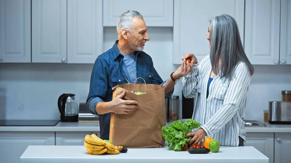 Senior man giving cherry tomatoes to asian wife while unpacking paper bag in kitchen — Stock Photo