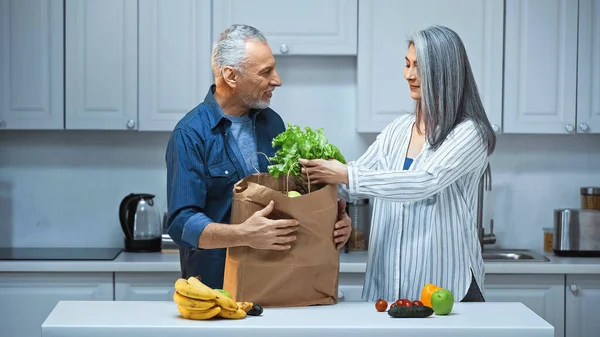 Idoso interracial casal desembalagem saco de papel com legumes frescos e frutas na cozinha — Fotografia de Stock