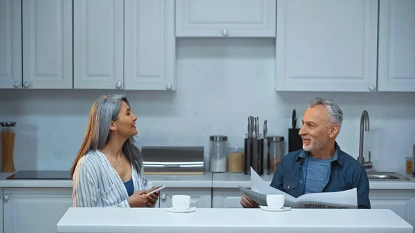 Elderly interracial couple talking during morning coffee in kitchen — Stock Photo