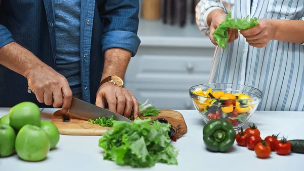 Abgeschnittene Ansicht eines älteren Mannes, der Salat schneidet, während er das Frühstück in der Nähe seiner Frau zubereitet — Stockfoto