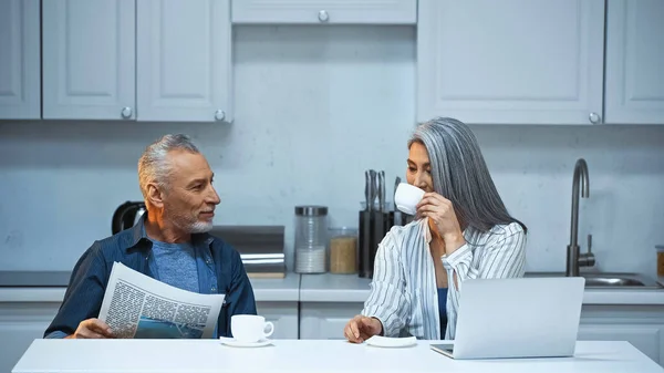 Gris pelo asiático mujer beber café cerca marido con periódico en cocina - foto de stock