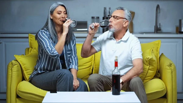 Grey haired interracial couple drinking red wine on sofa at home — Stock Photo
