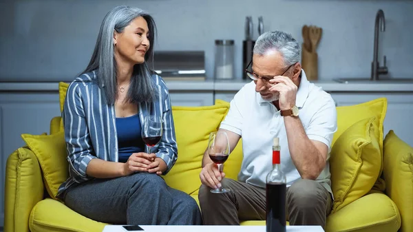 Feliz pareja interracial ancianos sosteniendo vasos con vino tinto en el sofá en casa - foto de stock