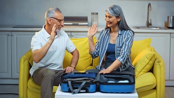 Happy elderly interracial couple giving high five near open travel bag — Stock Photo