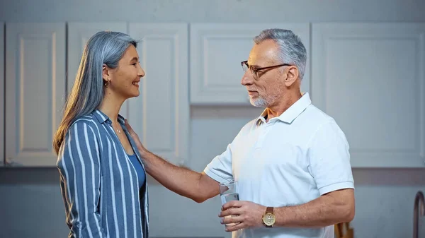 Side view of smiling senior man holding glass of water while touching shoulder of happy asian wife — Stock Photo