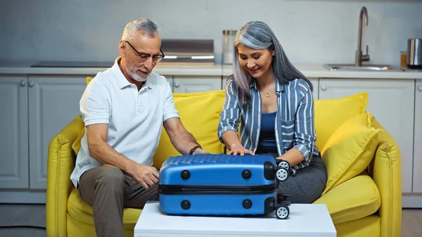 Senior interracial pareja sentado en sofá en casa cerca maleta - foto de stock
