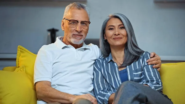 Happy grey haired man embracing smiling asian wife while sitting on sofa at home — Stock Photo