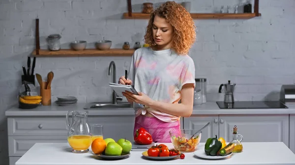 Curly woman with notebook weighing bell pepper near vegetables and orange juice on table — Stock Photo