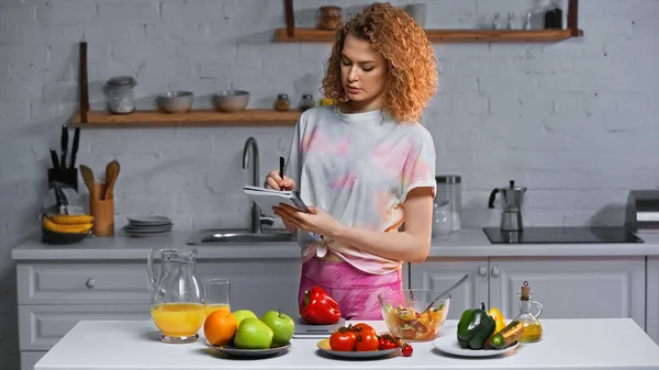 Mujer rizada con cuaderno que pesa pimiento cerca de verduras y jugo de naranja en la mesa - foto de stock