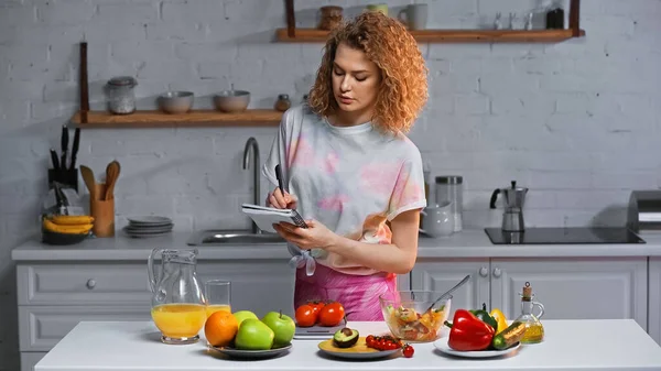 Woman writing in notebook and weighing tomatoes near vegetables and orange juice on table — Stock Photo